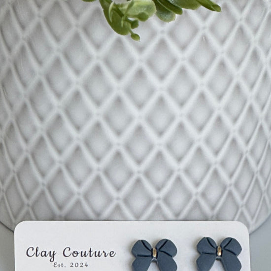 Slate Blue color Polymer Clay bow earrings on a Clay Couture card with a white textured background
