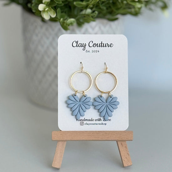 Leaf earrings  in Dusty Blue color made out of polymer clay with hypoallergenic attachments on a Clay Couture branded card with a plant in the background.