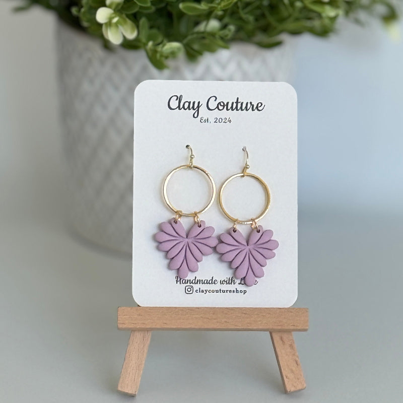 Leaf earrings  in Mauve color made out of polymer clay with hypoallergenic attachments on a Clay Couture branded card with a plant in the background.
