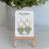 Leaf earrings  in Sage color made out of polymer clay with hypoallergenic attachments on a Clay Couture branded card with a plant in the background.