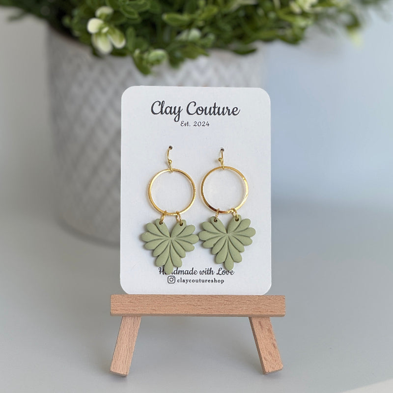 Leaf earrings  in Sage color made out of polymer clay with hypoallergenic attachments on a Clay Couture branded card with a plant in the background.
