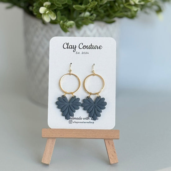 Leaf earrings  in Slate Blue color made out of polymer clay with hypoallergenic attachments on a Clay Couture branded card with a plant in the background.