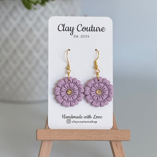 Mauve flower earrings on a Clay Couture branded card with a wooden stand.