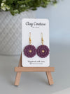 Plum flower earrings on a Clay Couture branded card with a wooden stand.