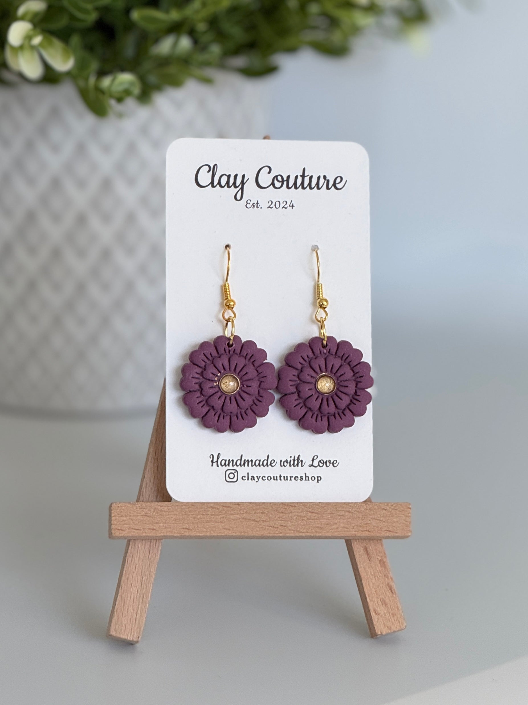Plum flower earrings on a Clay Couture branded card with a wooden stand.