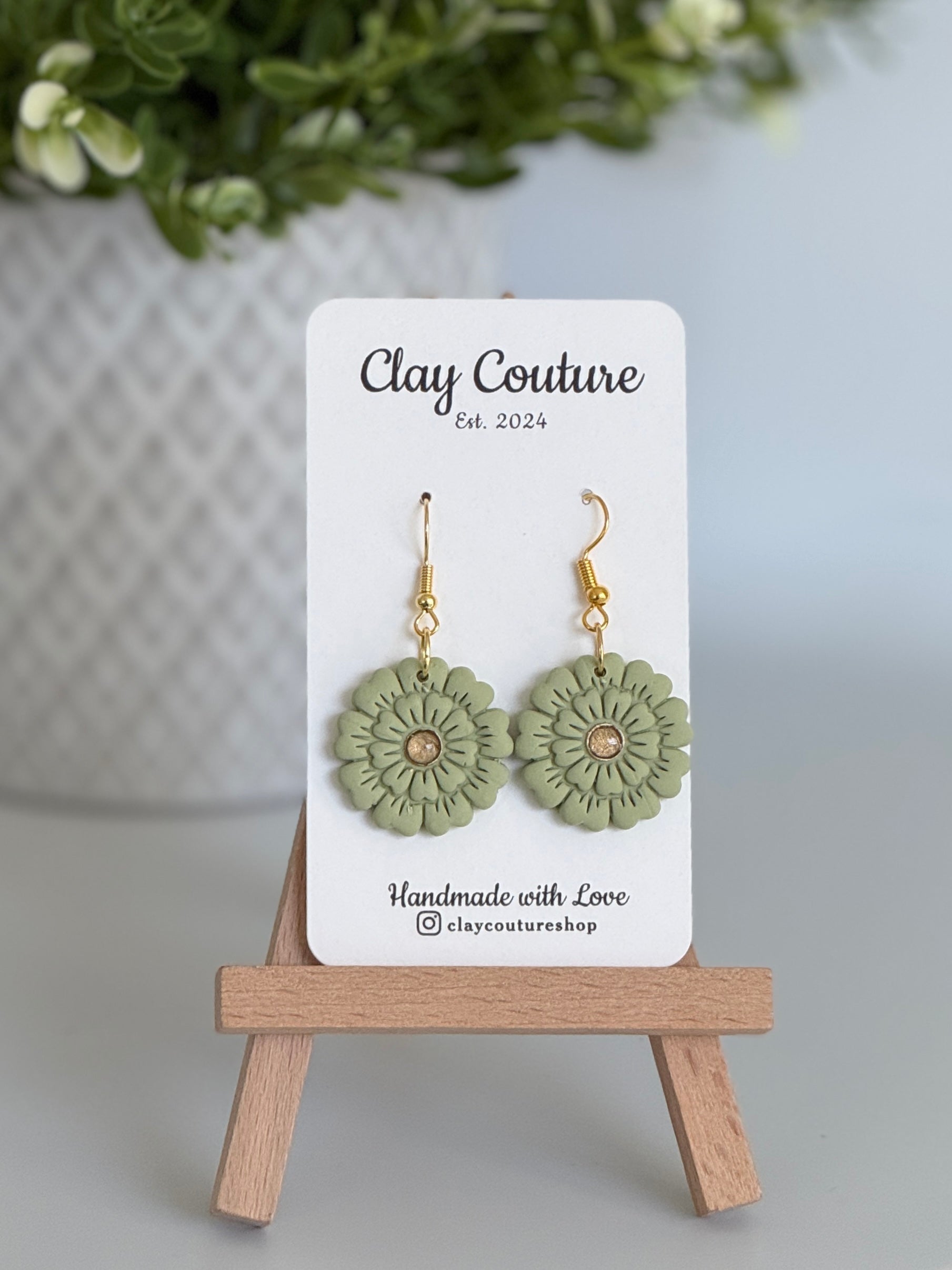 Sage flower earrings on a Clay Couture branded card with a wooden stand.