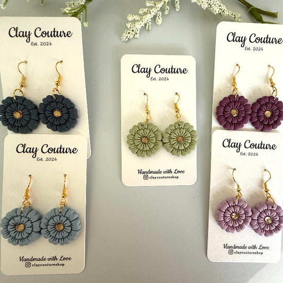 Set of flower-shaped earrings on 'Clay Couture' branded cards with a light gray background.