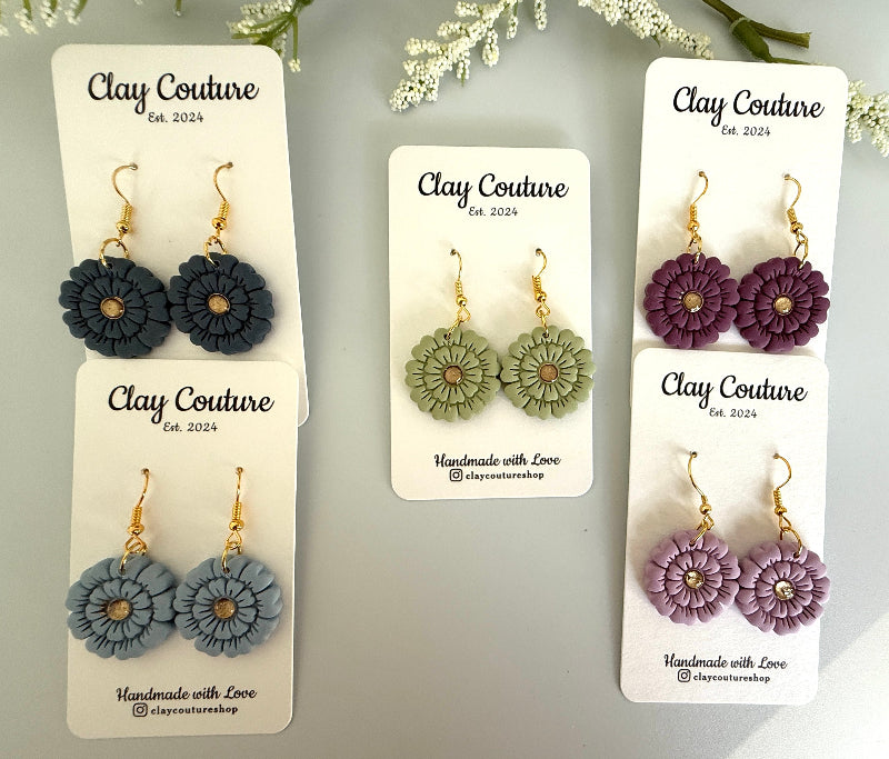 Set of flower-shaped earrings on 'Clay Couture' branded cards with a light gray background.