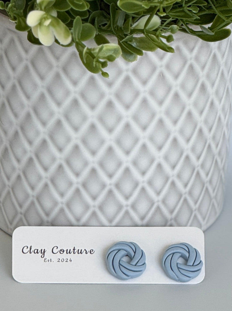 Dusty Blue Polymer Clay knot earrings on a Clay Couture card with a potted plant in the background