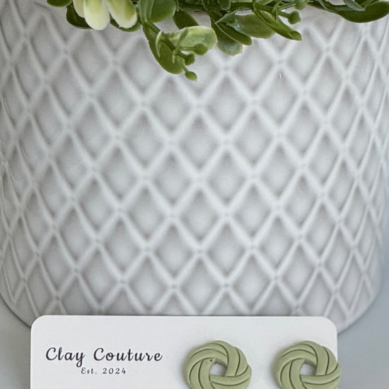 Sage Polymer Clay knot earrings on a Clay Couture card with a potted plant in the background