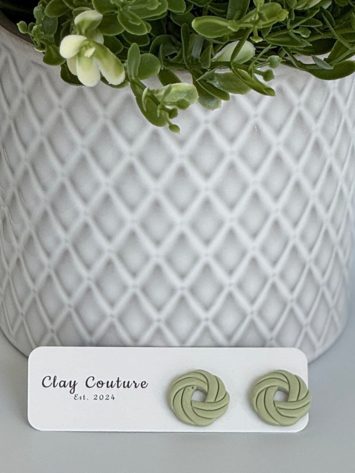 Sage Polymer Clay knot earrings on a Clay Couture card with a potted plant in the background