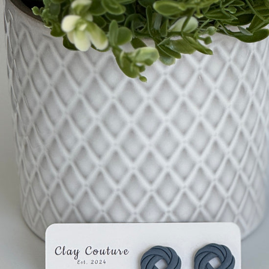 Slate Grey Polymer Clay knot earrings on a Clay Couture card with a potted plant in the backgroud.
