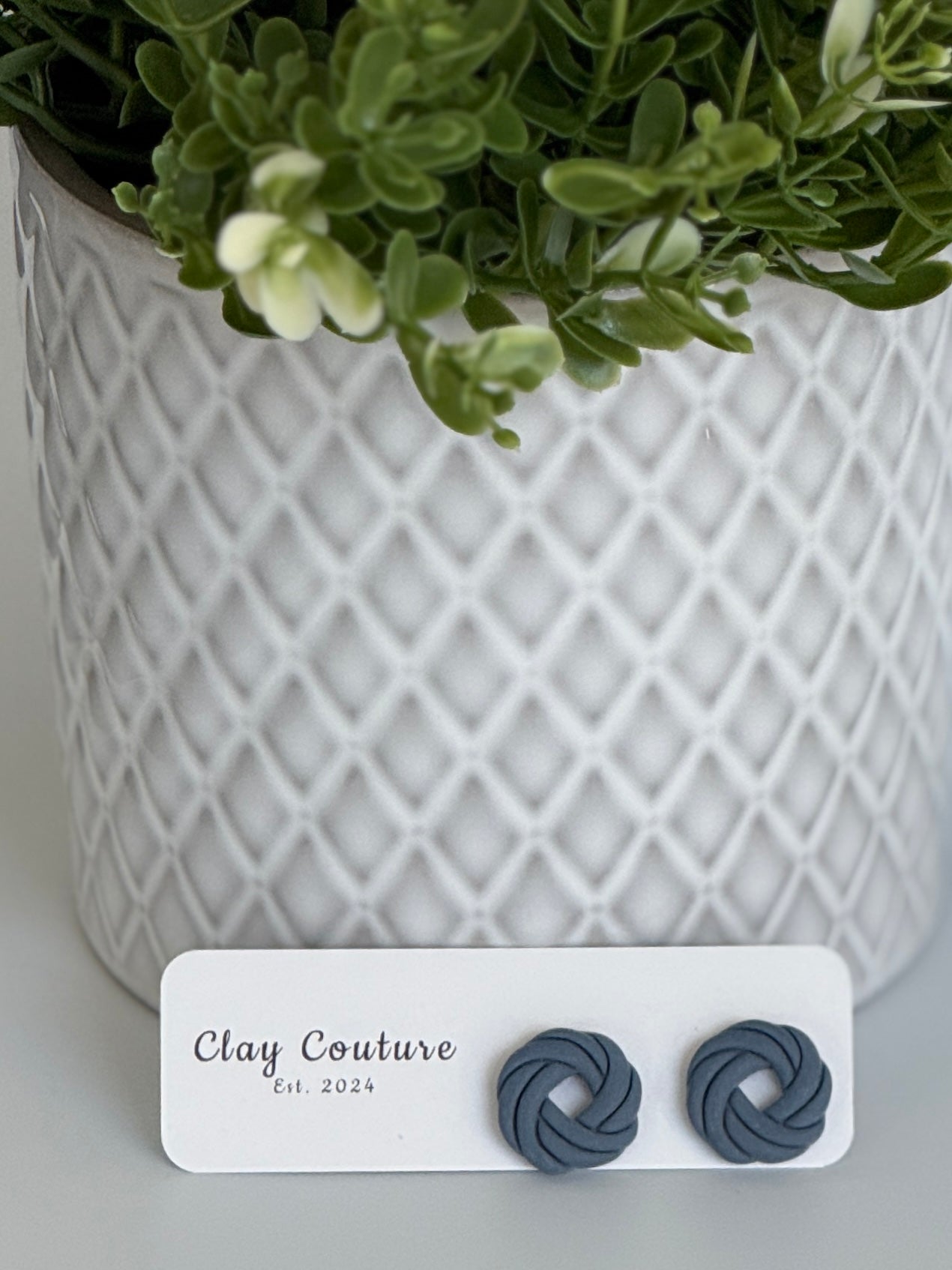 Slate Grey Polymer Clay knot earrings on a Clay Couture card with a potted plant in the backgroud.