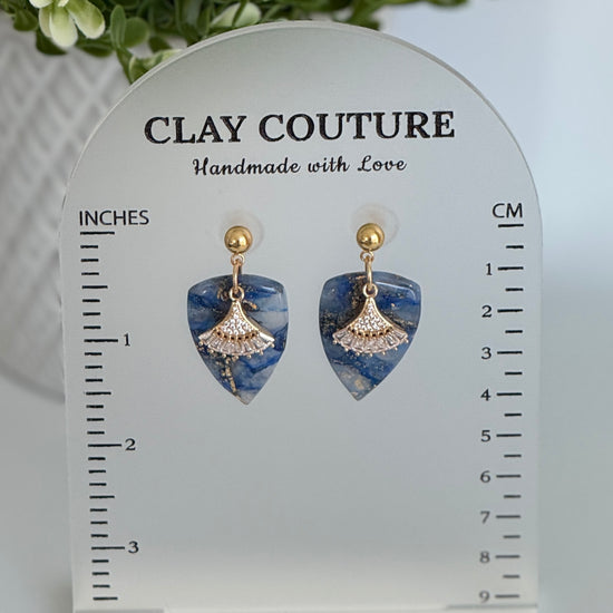 Sapphire Blue dagger shaped polymer clay  earrings  with a resin finish and gold accents on a Clay Couture branded measuring stand showing the length of the earrings
