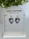 Sapphire Blue dagger shaped polymer clay  earrings  with a resin finish and gold accents on a Clay Couture branded measuring stand showing the length of the earrings