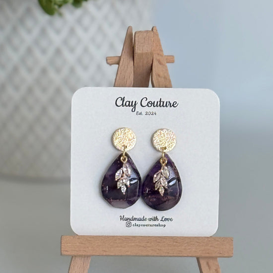 Amethyst purple teardrop shaped polymer clay  earrings  with a resin finish and gold accents on a Clay Couture branded card with a wooden stand.