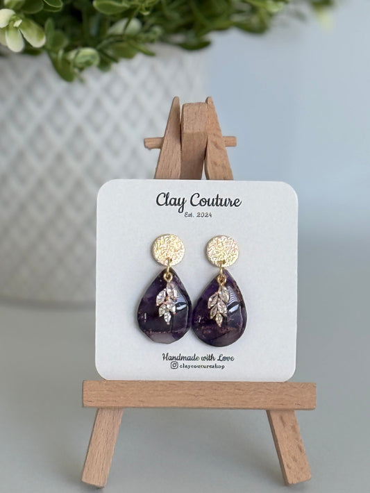 Amethyst purple teardrop shaped polymer clay  earrings  with a resin finish and gold accents on a Clay Couture branded card with a wooden stand.