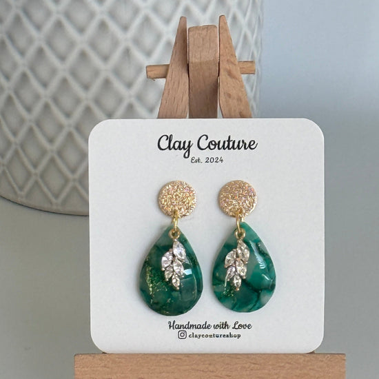 Emerald green teardrop shaped polymer clay  earrings  with a resin finish and gold accents on a Clay Couture branded card with a wooden stand.