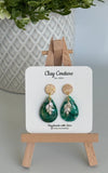 Emerald green teardrop shaped polymer clay  earrings  with a resin finish and gold accents on a Clay Couture branded card with a wooden stand.
