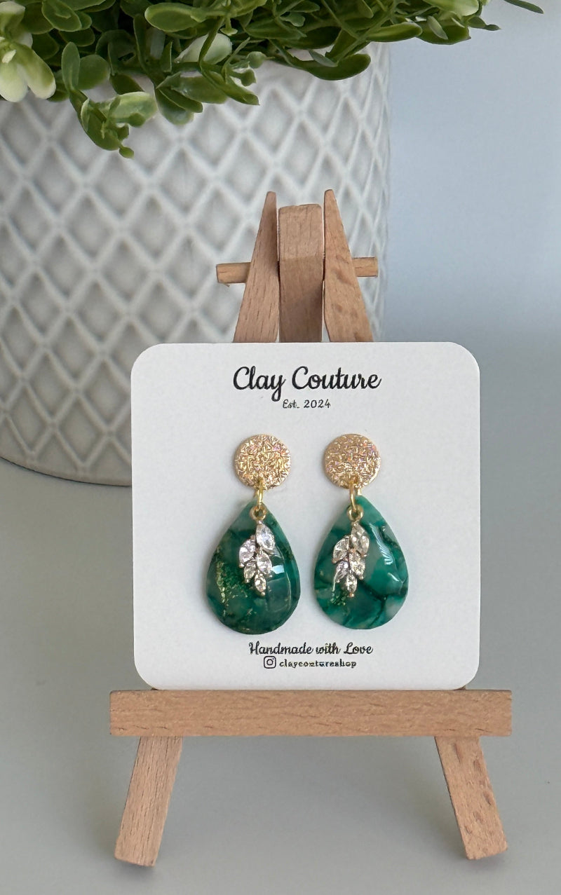 Emerald green teardrop shaped polymer clay  earrings  with a resin finish and gold accents on a Clay Couture branded card with a wooden stand.
