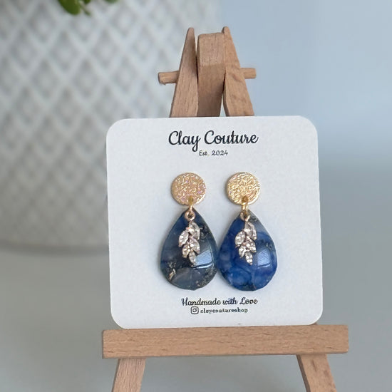 Sapphire Blue teardrop shaped polymer clay  earrings  with a resin finish and gold accents on a Clay Couture branded card with a wooden stand.