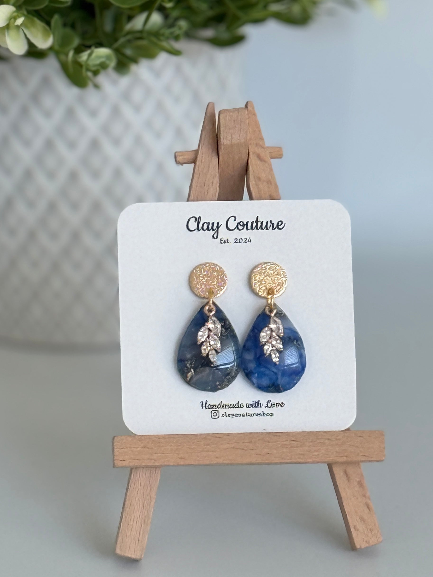 Sapphire Blue teardrop shaped polymer clay  earrings  with a resin finish and gold accents on a Clay Couture branded card with a wooden stand.