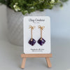 Amethyst purple earrings  with a gold chain and bow stud on a 'Clay Couture' branded card with a plant in the background