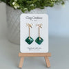 Emerald Green earrings  with a gold chain and bow stud on a 'Clay Couture' branded card with a plant in the background