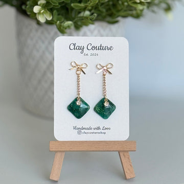 Emerald Green earrings  with a gold chain and bow stud on a 'Clay Couture' branded card with a plant in the background