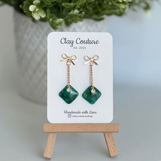 Emerald Green earrings  with a gold chain and bow stud on a 'Clay Couture' branded card with a plant in the background