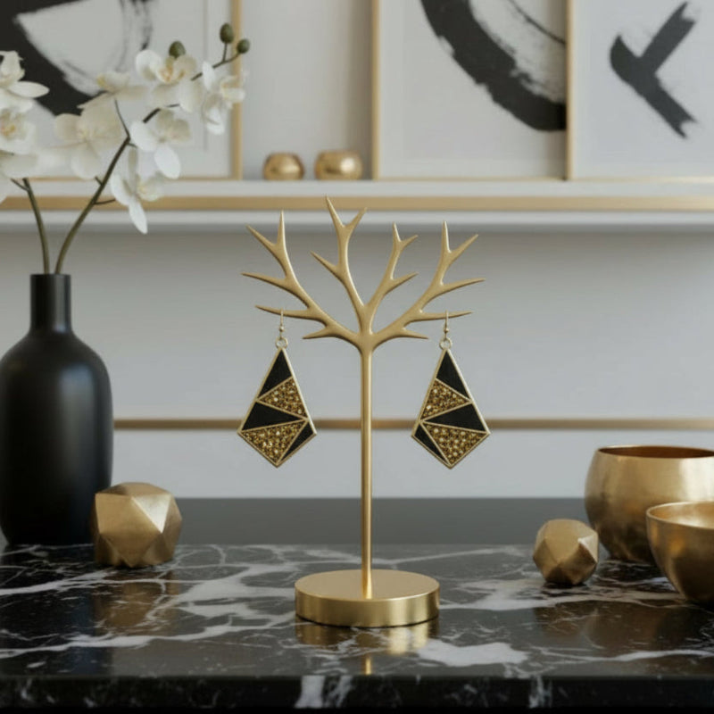 Gold earring stand with geometric earrings on a marble surface, with decorative items in the background.